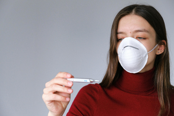 Von Long Covid betroffene Frau mit Maske prüft Fieberthermometer 